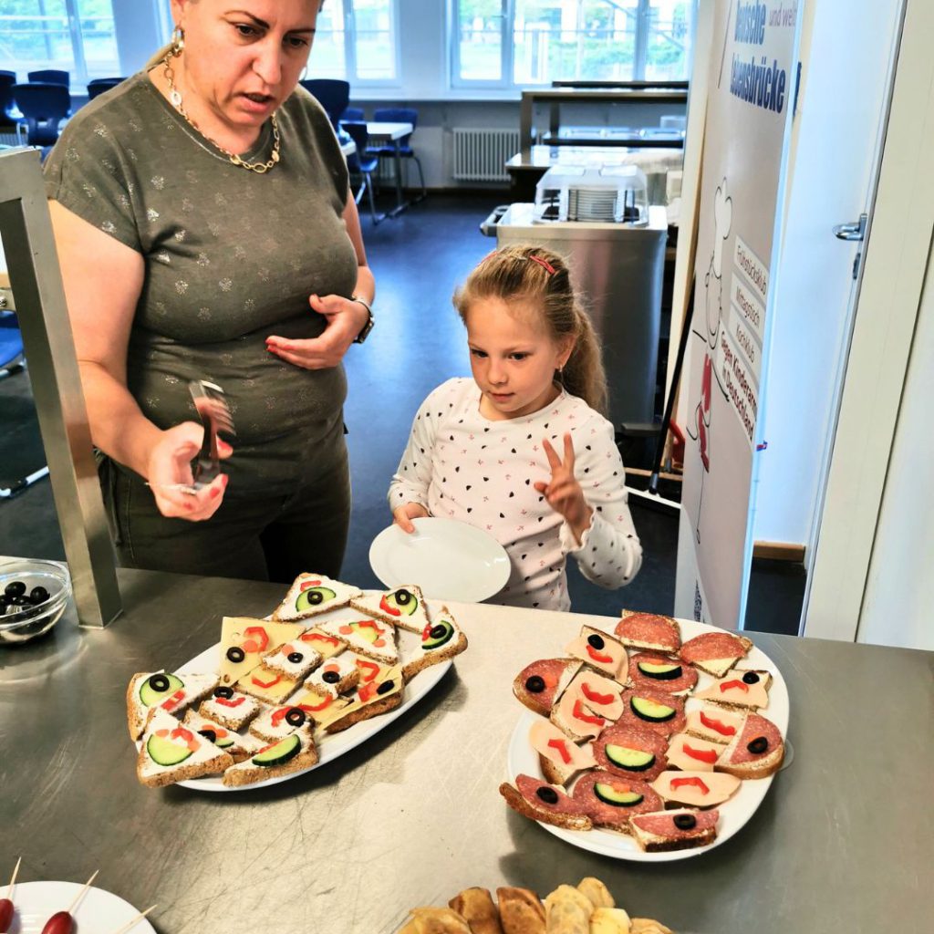 Betreuerin und Kind bereiten im Frühstücksklub der Deutschen Lebensbrücke e.V. in München-Obersendling kreativ belegte Brote für das gemeinsame Frühstück vor.