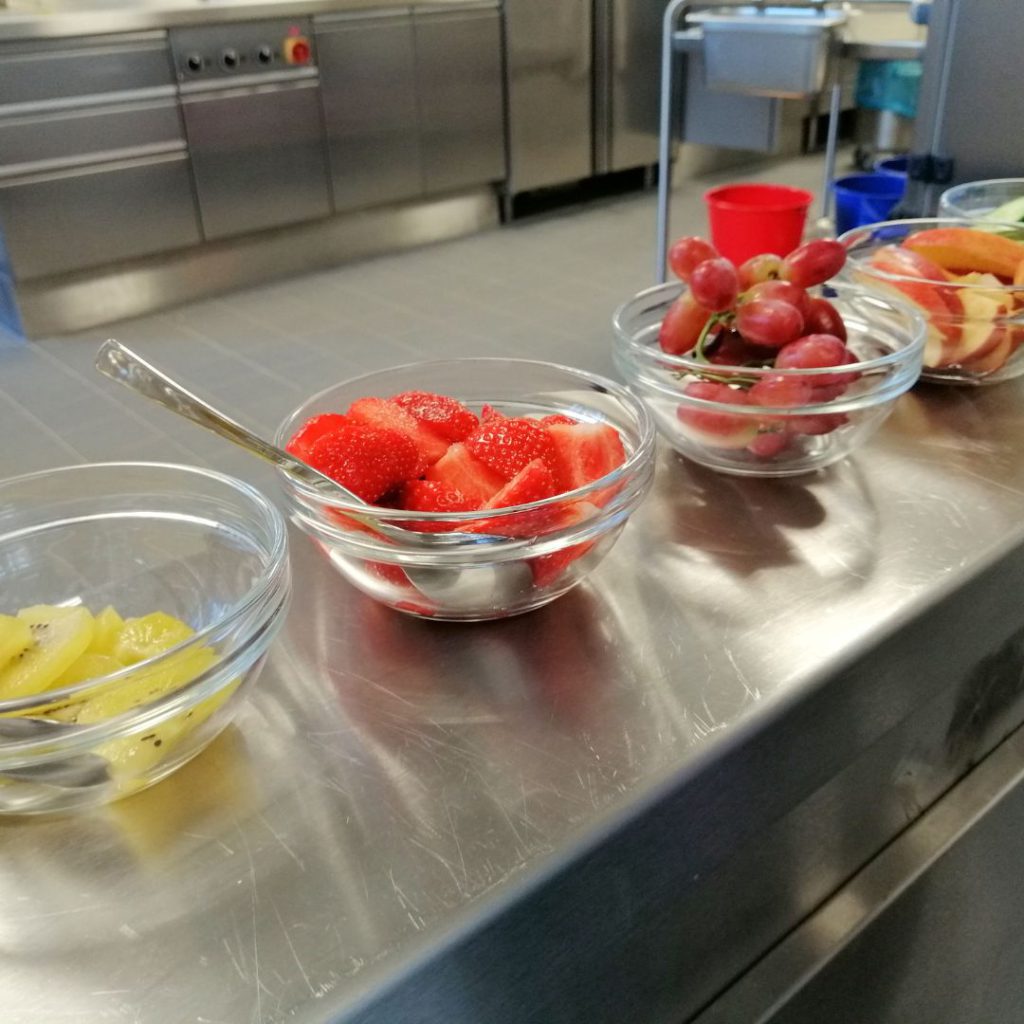 Nahaufnahme von Glasschalen voller Kiwi, Erdbeeren, Trauben und Äpfel. Hier erhalten die Kinder frisch belegte Brote, frisches Gemüse und saftiges Obst – auf jeden Fall ausgewogen.
