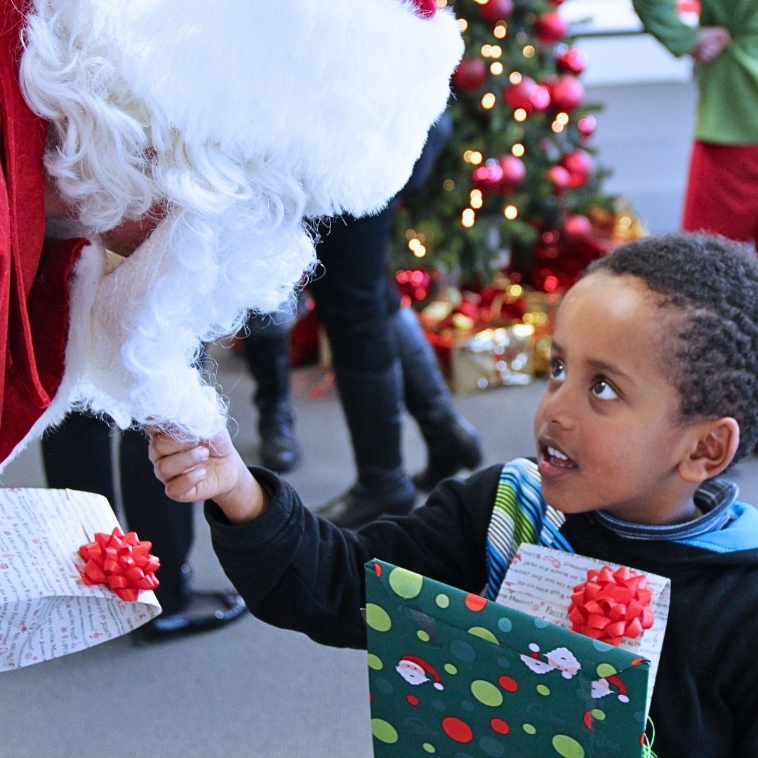 Zur Weihnachtszeit für Kinder spenden: Kleiner afrikanischer Junge erhält sein Weihnachtsgeschenk und zieht dem Weihnachtsmann lachend am Bart.